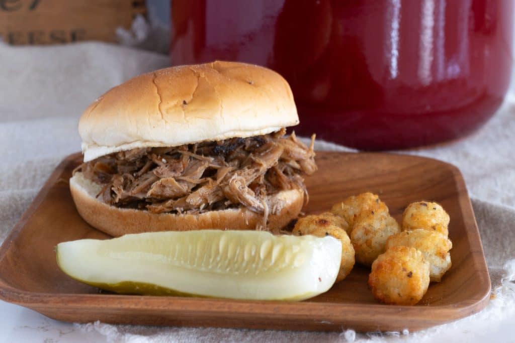 A barbecued pulled pork sandwich with a pot of pulled pork in the background.