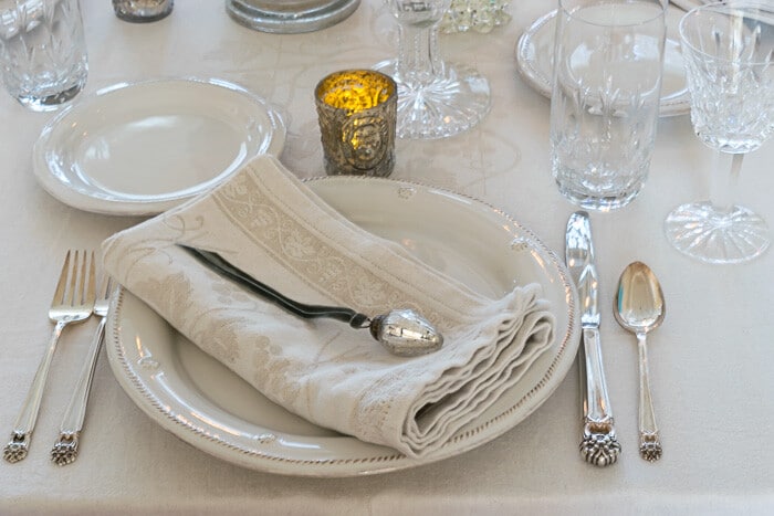 Winter White Table setting features white plates, neutral beige napkins, silver, crystal, and a mercury glass ornament.