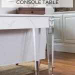 console table with acrylic legs and a faux marble top with white sofa in the background.