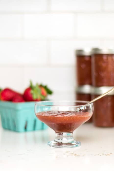 Bowl of strawberry rhubarb jam with jars of jam and a container of strawberries in the background.