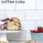 Strawberry Coffee Cake on a Pedestal in front of a white tile wall, with a basket of strawberries in the background.