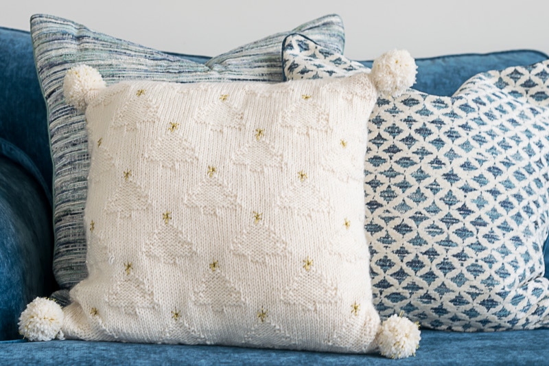 White knit pillow with pom poms against a blue sofa and blue and white pillows.
