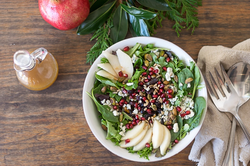 Overhead image of Christmas Salad in a white bowl on a wood surface with greens and a pomegranate in the background.
