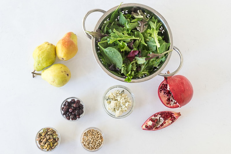 Overhead shot of ingredients for Festive Christmas Salad, including mixed greens, pears, dried cranberries, pistachios, sunflower seeds, pomegranate and gorgonzola cheese.