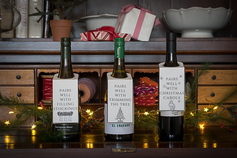 Three bottles of wine with Christmas bottle neck tags, on a hutch with small lights.