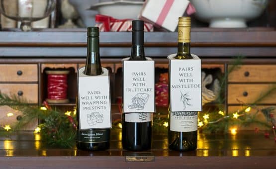 Three bottles of wine with Christmas wine bottle tags, on a hutch with small lights.