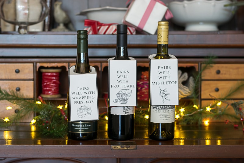 Three bottles of wine with Christmas wine bottle tags, on a hutch with small lights.