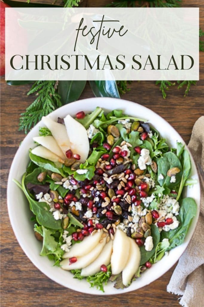 Overhead shot of Christmas Salad in a white bowl on a wood surface with greens and pomegranate in the background.