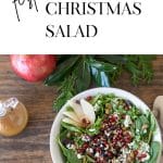 Overhead shot of Christmas Salad in a white bowl on a wood surface with greens and pomegranate in the background.