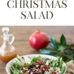 Image of Christmas Salad in a white bowl with a pomegranate, a jar of dressing and greens in the background.