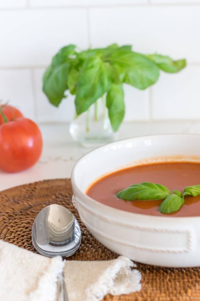 Bowl of healthy homemade tomato soup with fresh basil and tomatoes in the background.