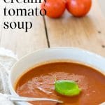 Bowl of healthy homemade tomato soup with fresh basil and tomatoes in the background.