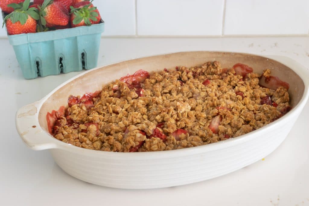 Strawberry Crisp recipe baked in a oval baking dish, with a container of strawberries in the background.