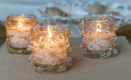 Three beaded DIY Macramé Wire Votive Holders filled with burning candles sitting on a book.