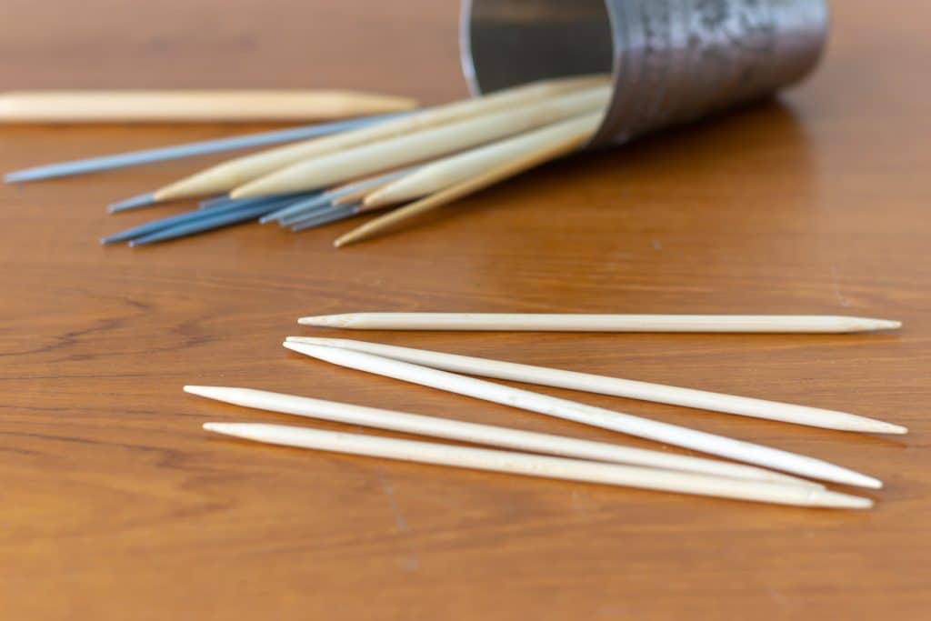 Double-pointed needles on a table, with a cup of double-pointed spilled in the background.