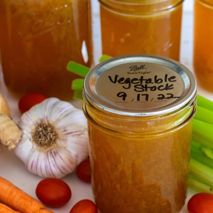 A jar of homemade vegetable stock, with other jars in the background and fresh vegetables scattered around.