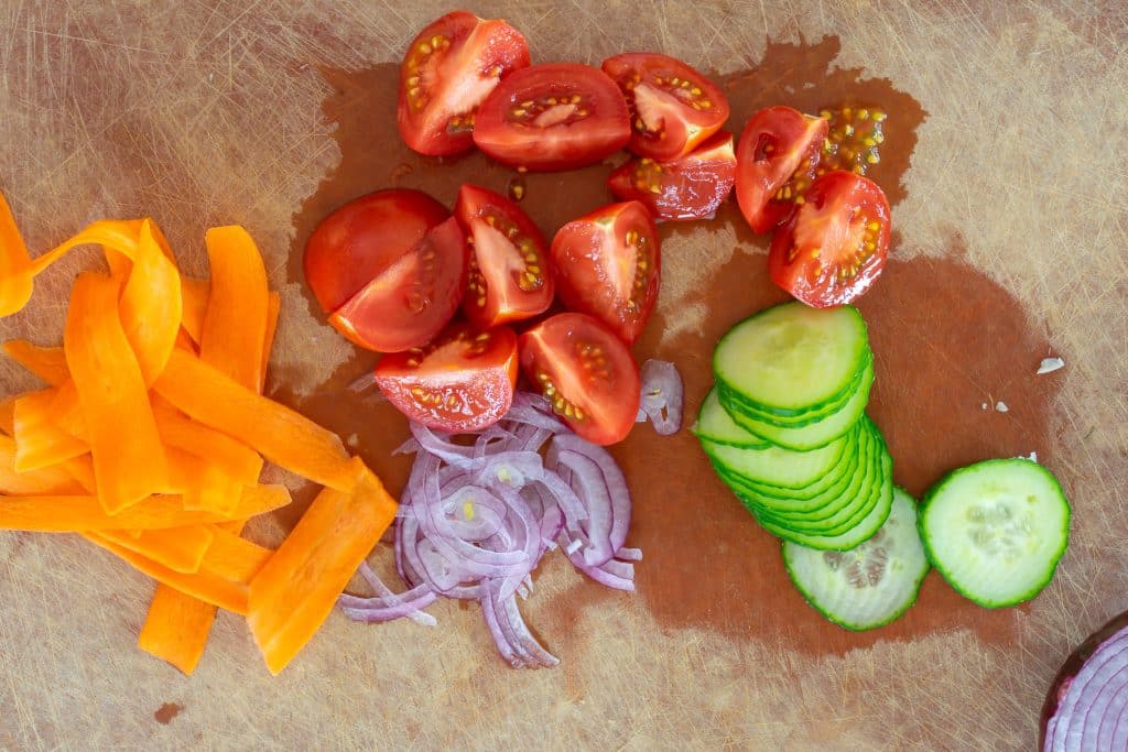Tomatoes, cucumbers, carrots, and onions sliced on a cutting board.
