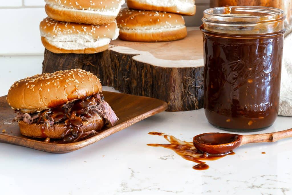 A sandwich sitting on a wooden plate with a jar of barbecue sauce.