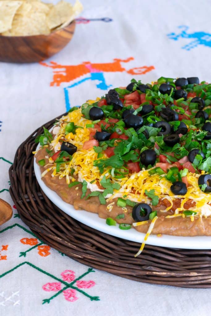 Mexican Layer Dip on a brown rattan platter on an embroidered tablecloth.