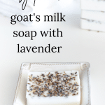 A bar of goat milk soap in a square soap dish with white washcloths and a stack of soap bars in the background.