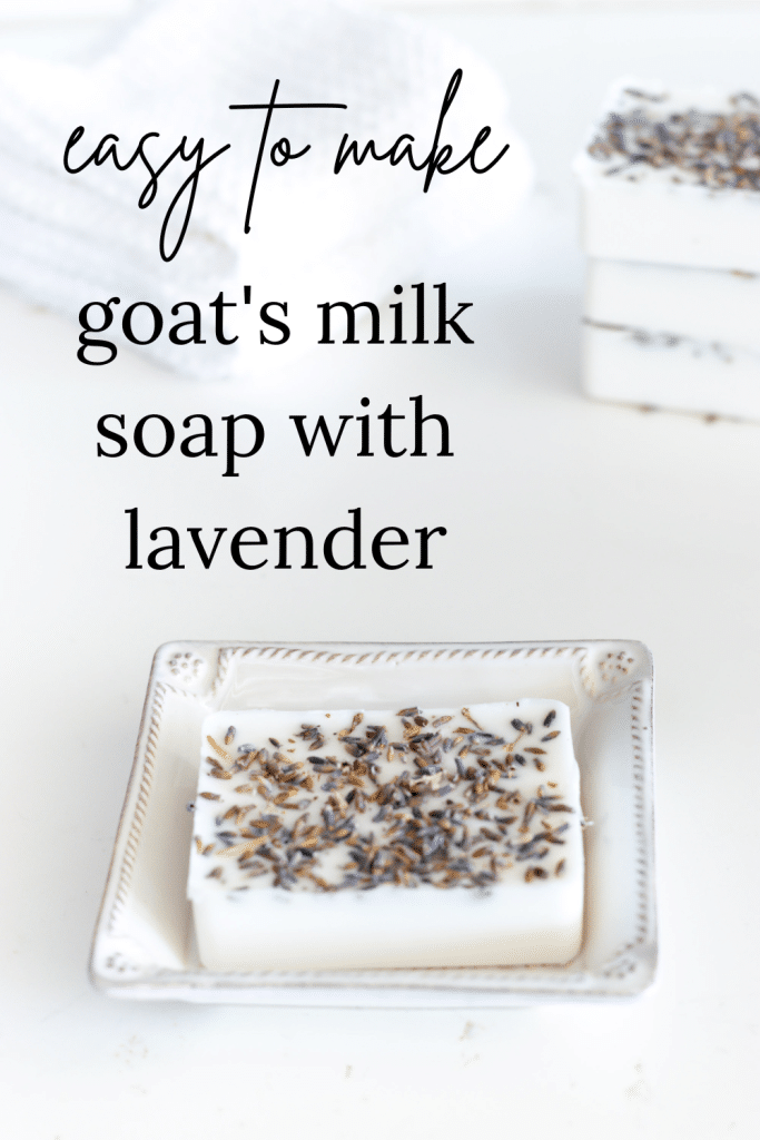 A bar of goat milk soap in a square soap dish with white washcloths and a stack of soap bars in the background.