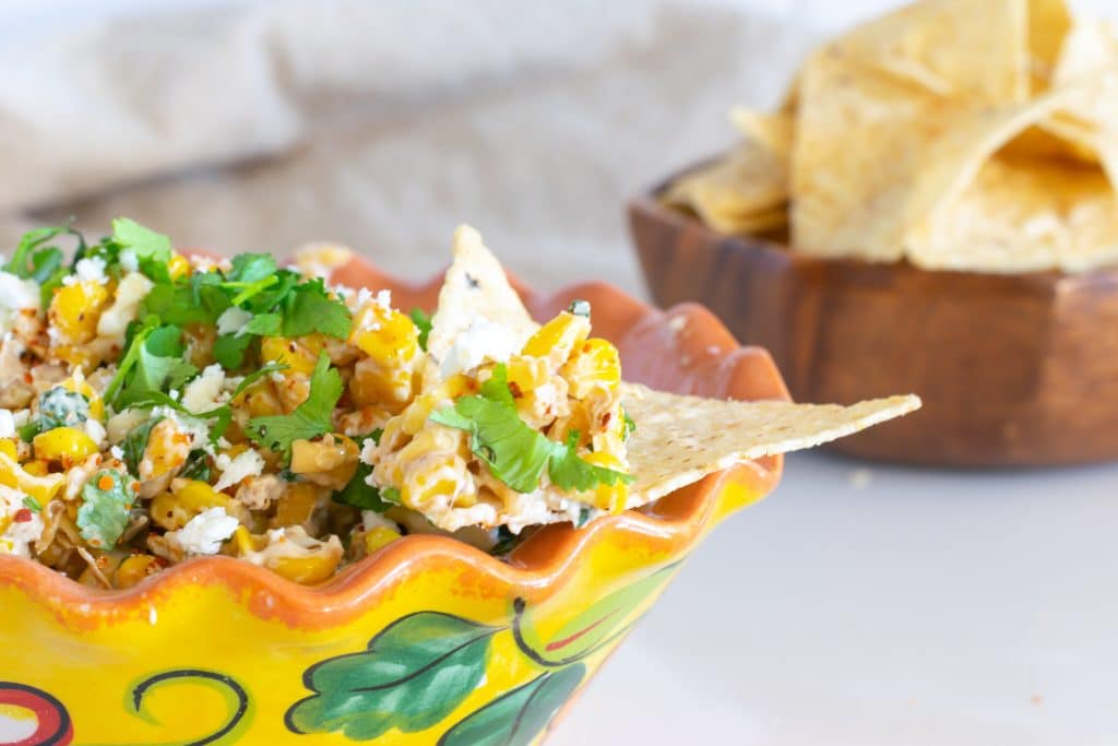 Elote dip in a yellow Mexican bowl with a chip for dipping.