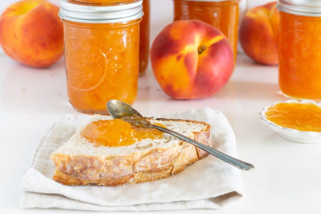 Peach preserves on a piece of bread with jars of preserves in the background.